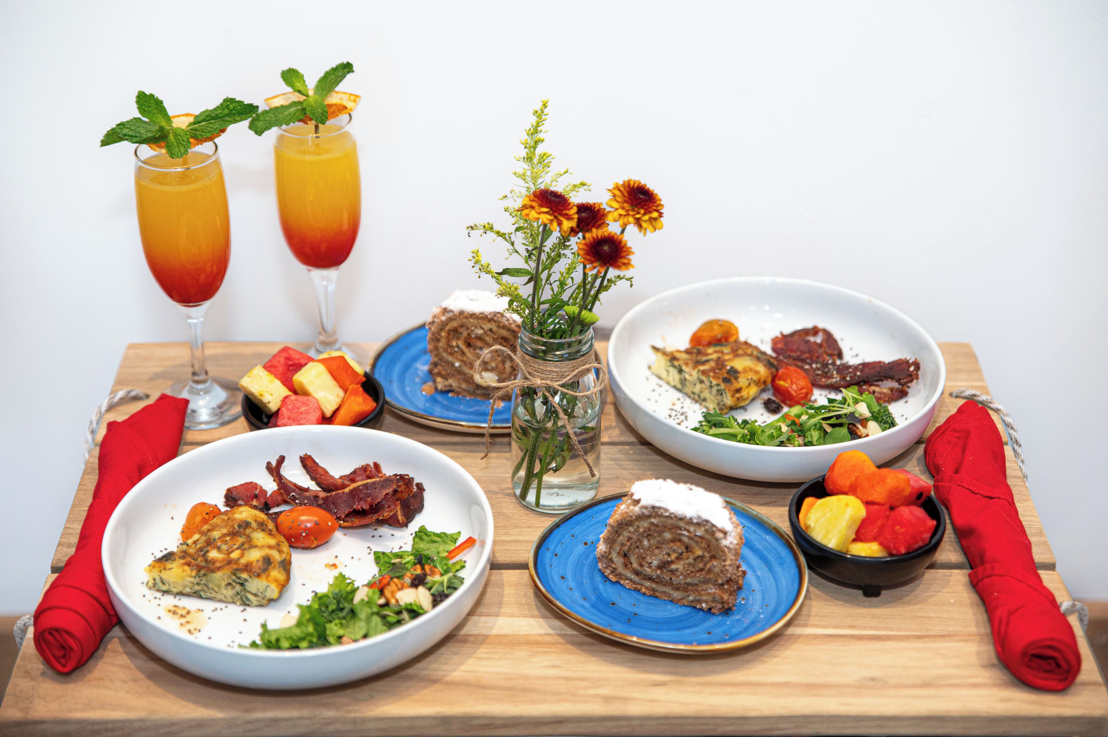 A table set with various dishes, including a salad, a rolled meat dish, and a slice of cake, along with two drinks and a vase of flowers.