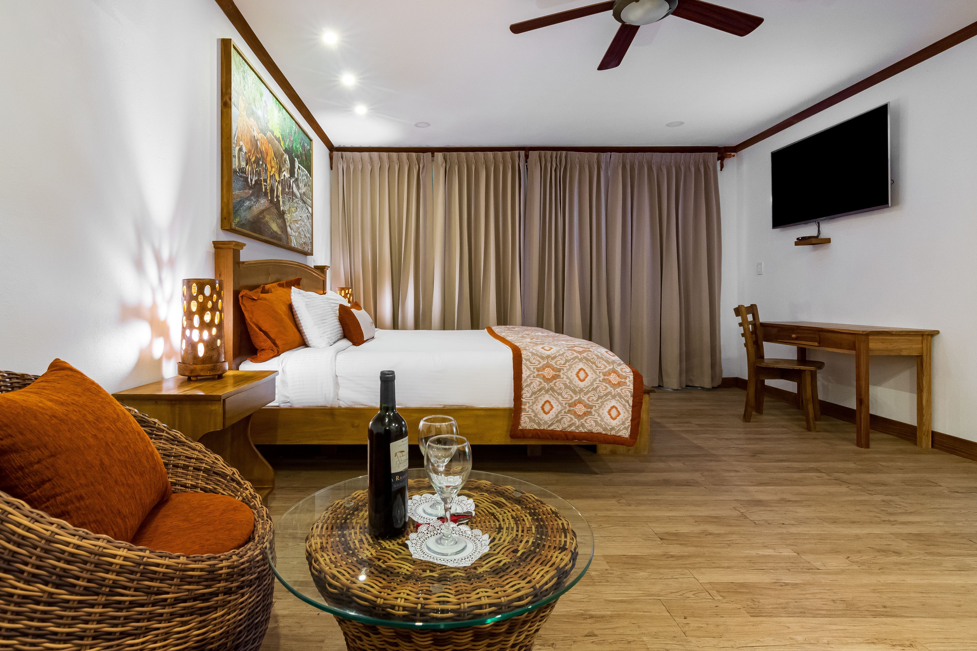 A hotel room with a bed, a wicker chair, a glass coffee table with a bottle and glasses, a desk, and a TV on the wall.
