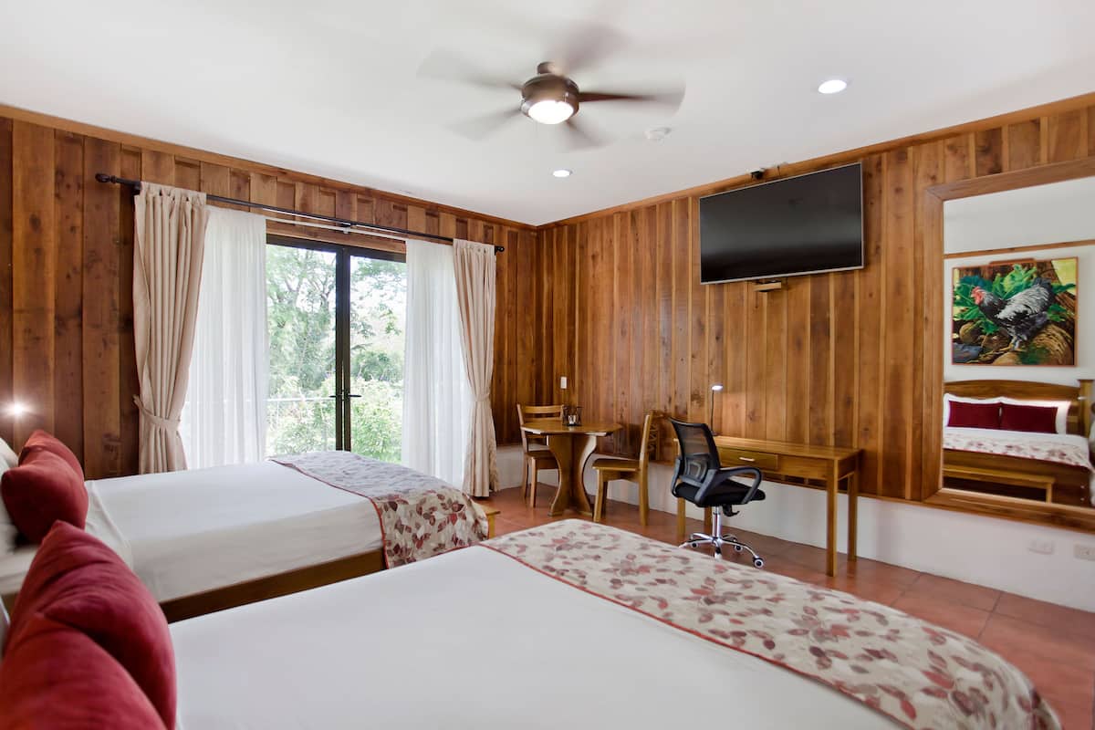 A hotel room with two beds, a desk, a chair, a television, and a ceiling fan.