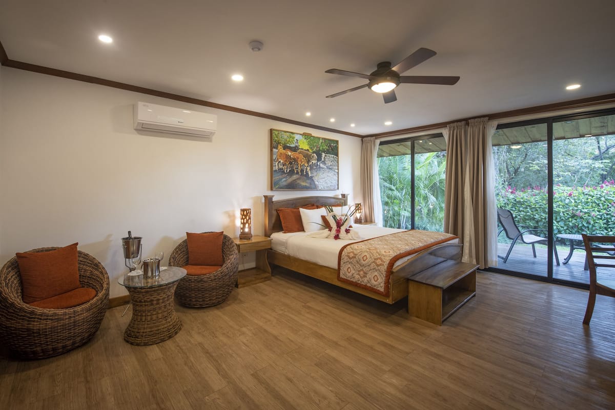 A spacious bedroom with a large bed, two wicker chairs, a small table, and a ceiling fan.