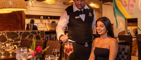 A restaurant setting with a waiter preparing a drink at a table.