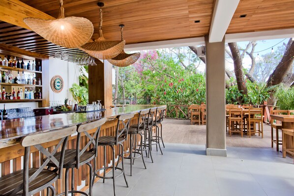 A bar area with a wooden bar counter, multiple stools, and hanging wicker lamps. - Hotel Hacienda Guachipelin (Curubandé)