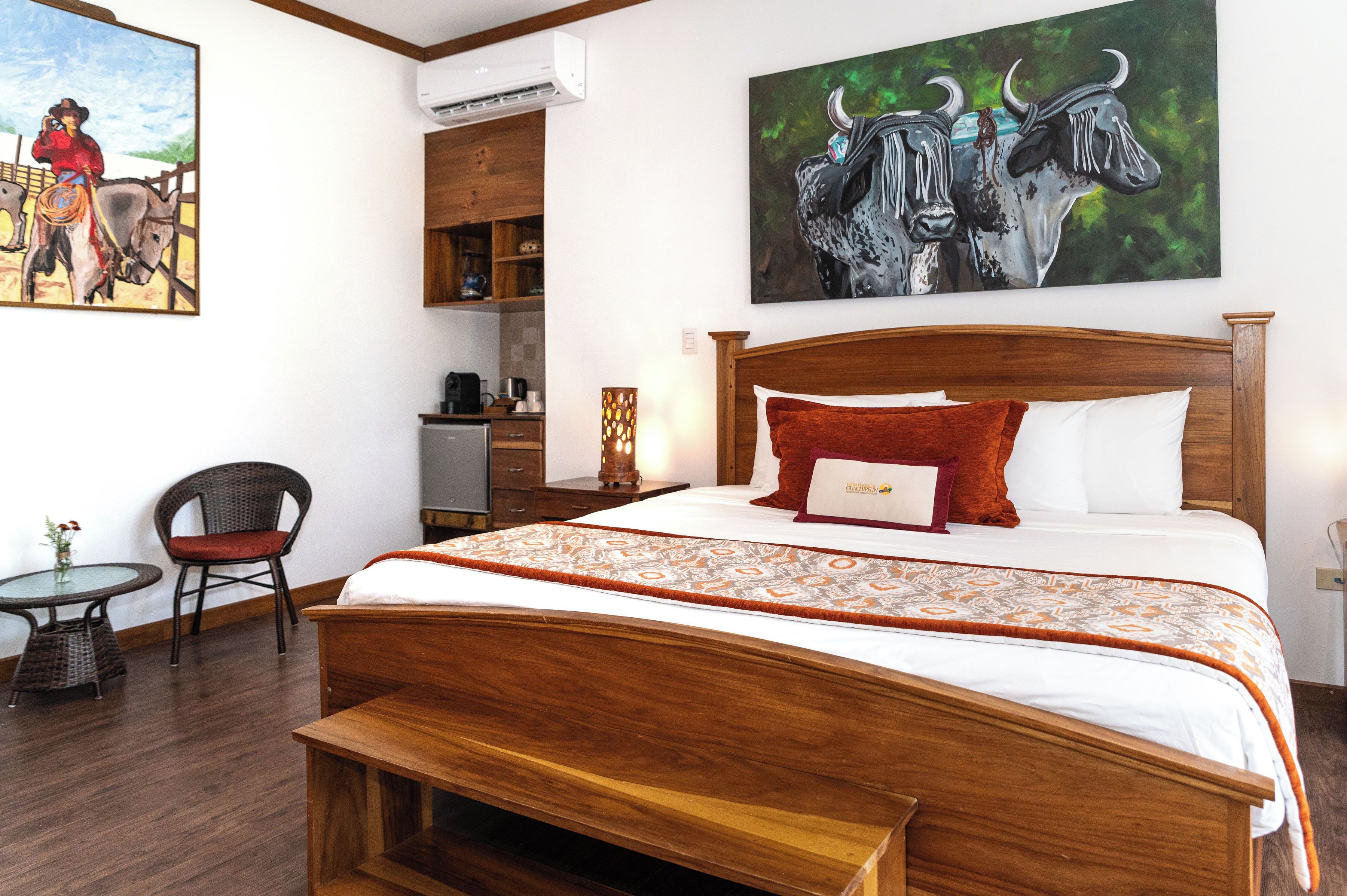A hotel room with a large bed, a wooden headboard, a small table with a chair, and a painting of bulls on the wall.