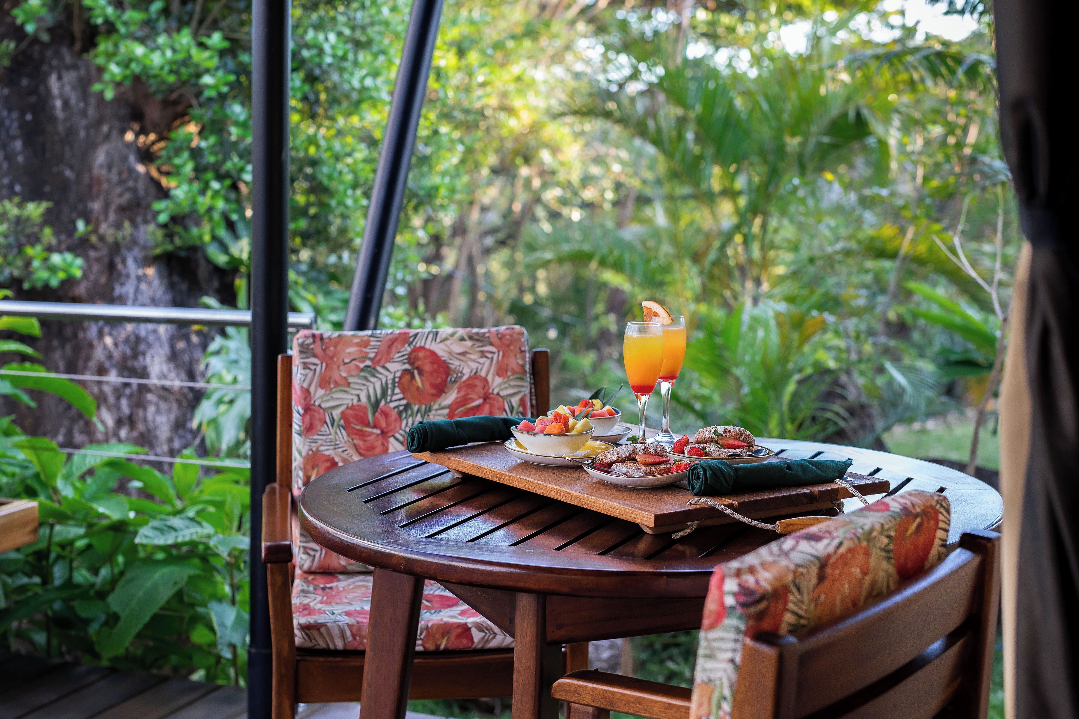A round wooden table with a floral cushion, a bowl of fruit, a glass of orange juice, and a napkin.