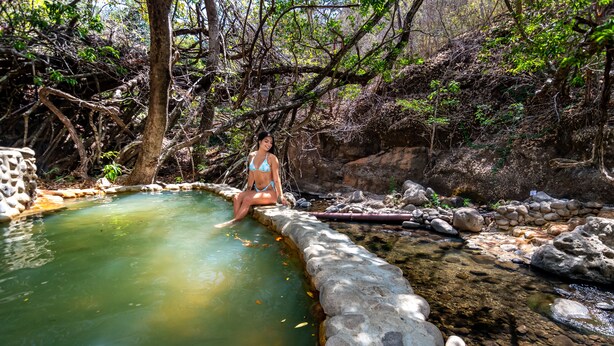 Une personne assise au bord d’une étendue d’eau claire, entourée de rochers et d’arbres.