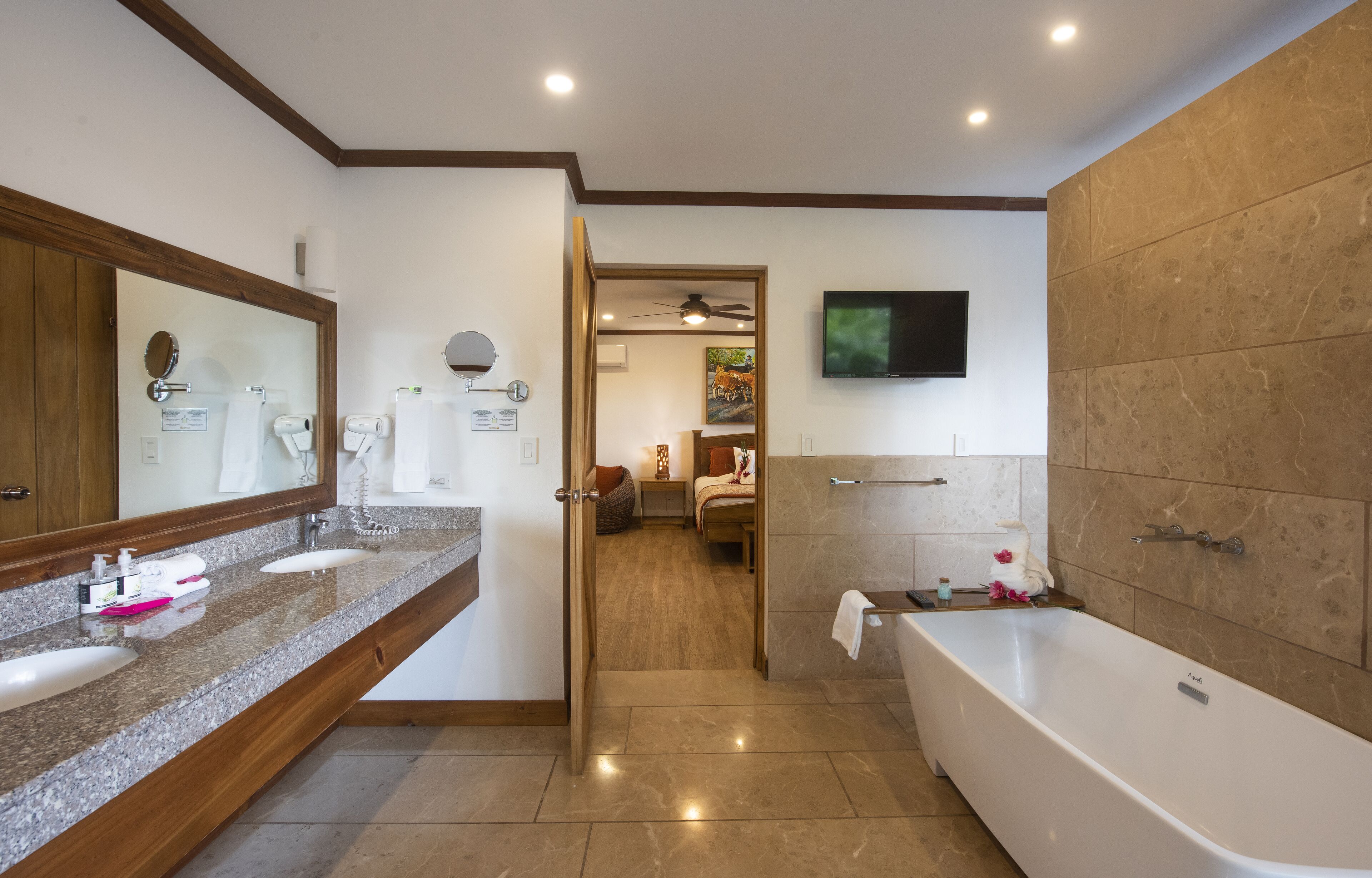 Un baño moderno con una bañera grande, dos lavamanos, un tocador, un espejo, un ventilador de techo y un televisor montado en la pared.