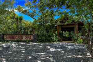 A sign for 'Simbiosis Spa' with a gravel path leading to a covered entrance surrounded by lush greenery.