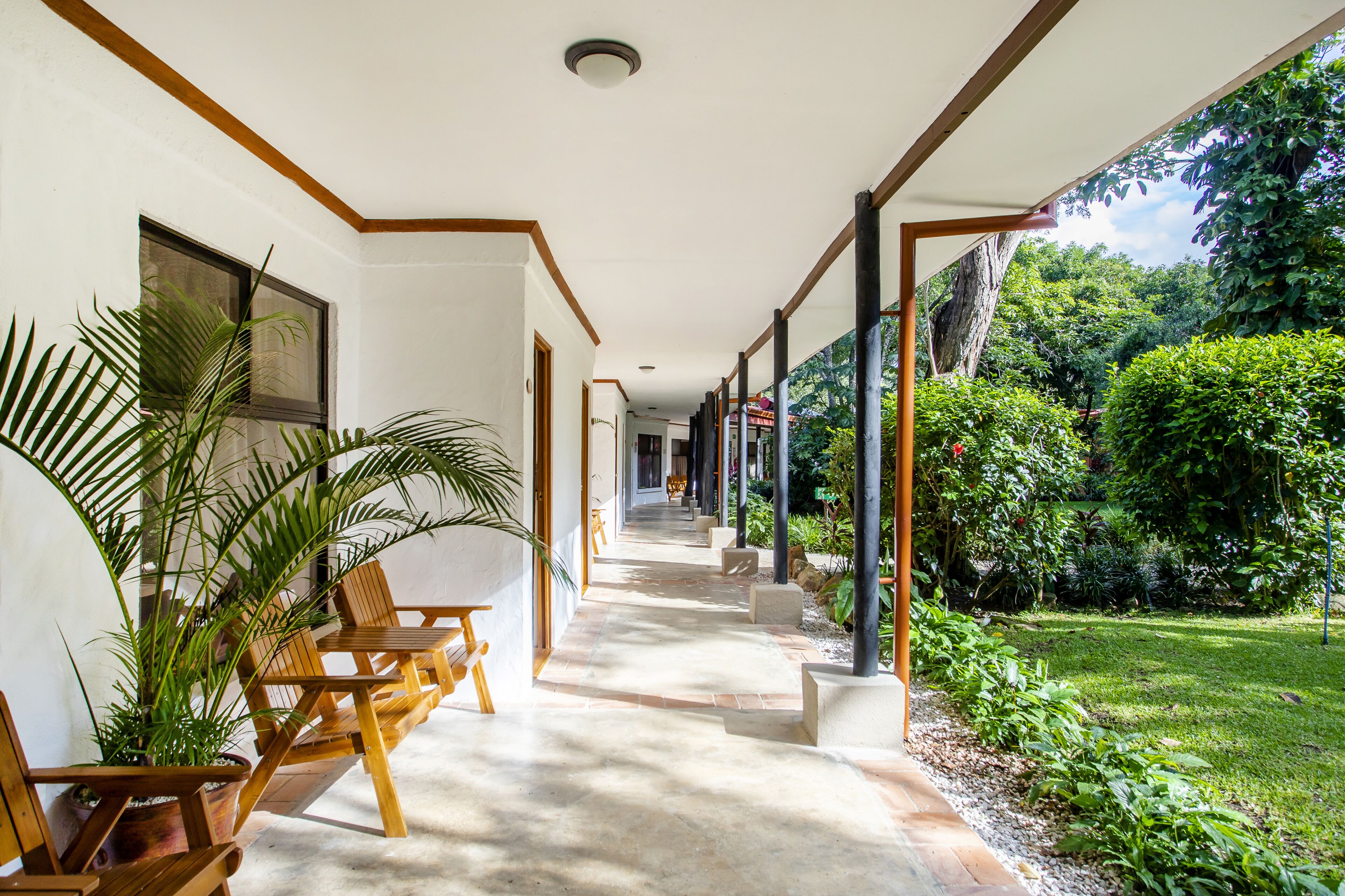 A covered outdoor corridor with wooden chairs and potted plants, leading to a lush garden area.