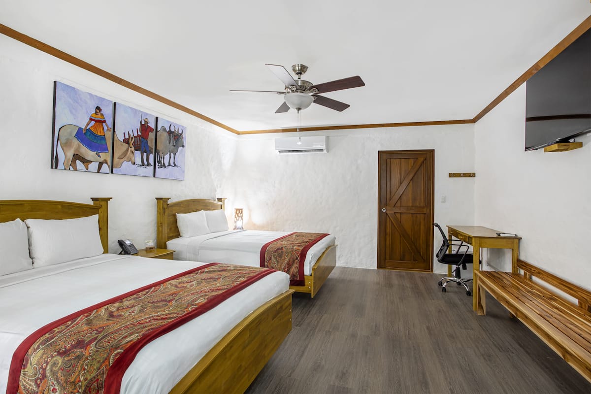 A hotel room with two beds, a ceiling fan, an air conditioning unit, a wooden desk, a chair, and a TV.