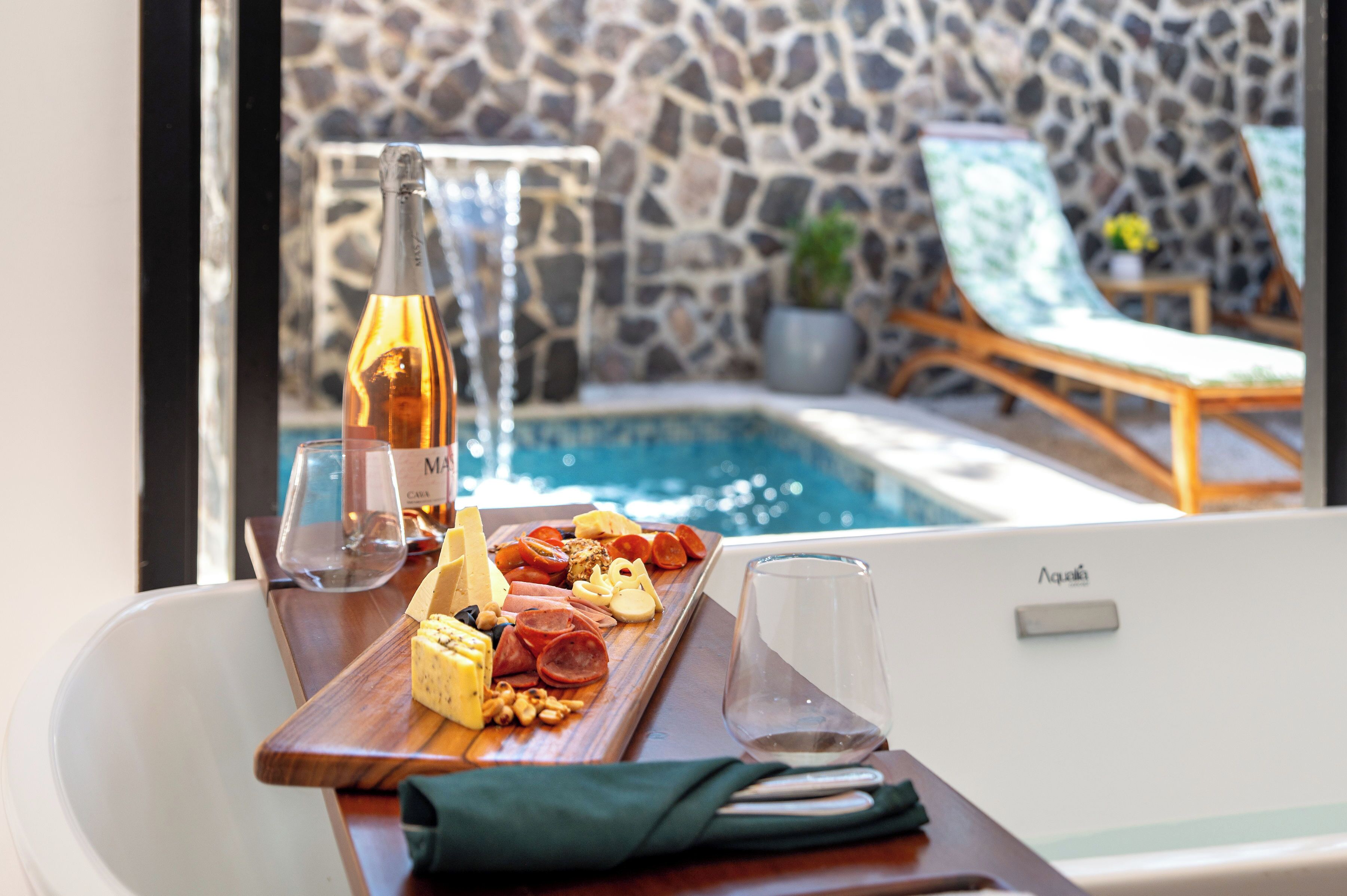 A poolside setting with a wooden tray of cheeses, meats, and a bottle of wine.