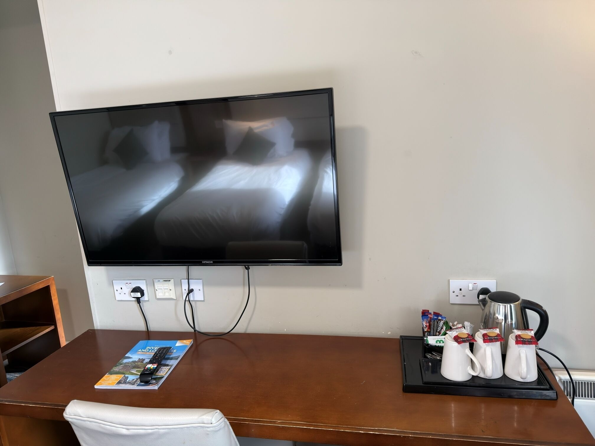 A hotel room with a flat-screen TV mounted on the wall, a wooden desk with a magazine, a white chair, and a tray with coffee mugs and a kettle.