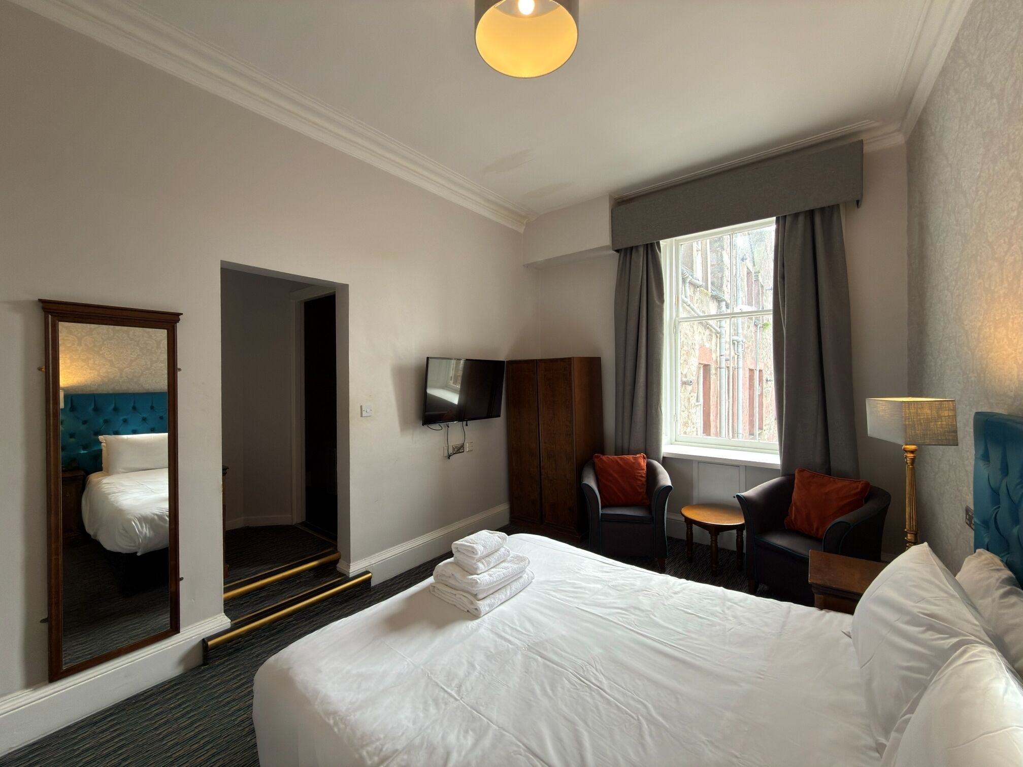A hotel room with a large bed, a television mounted on the wall, a seating area with two chairs, and a window with curtains.