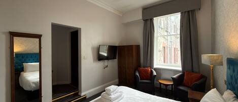 A hotel room with a large bed, a television mounted on the wall, a seating area with two chairs, and a window with curtains.