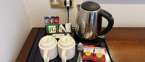 A coffee station with a kettle, two mugs, a spoon, and packets of coffee creamer and biscuits.