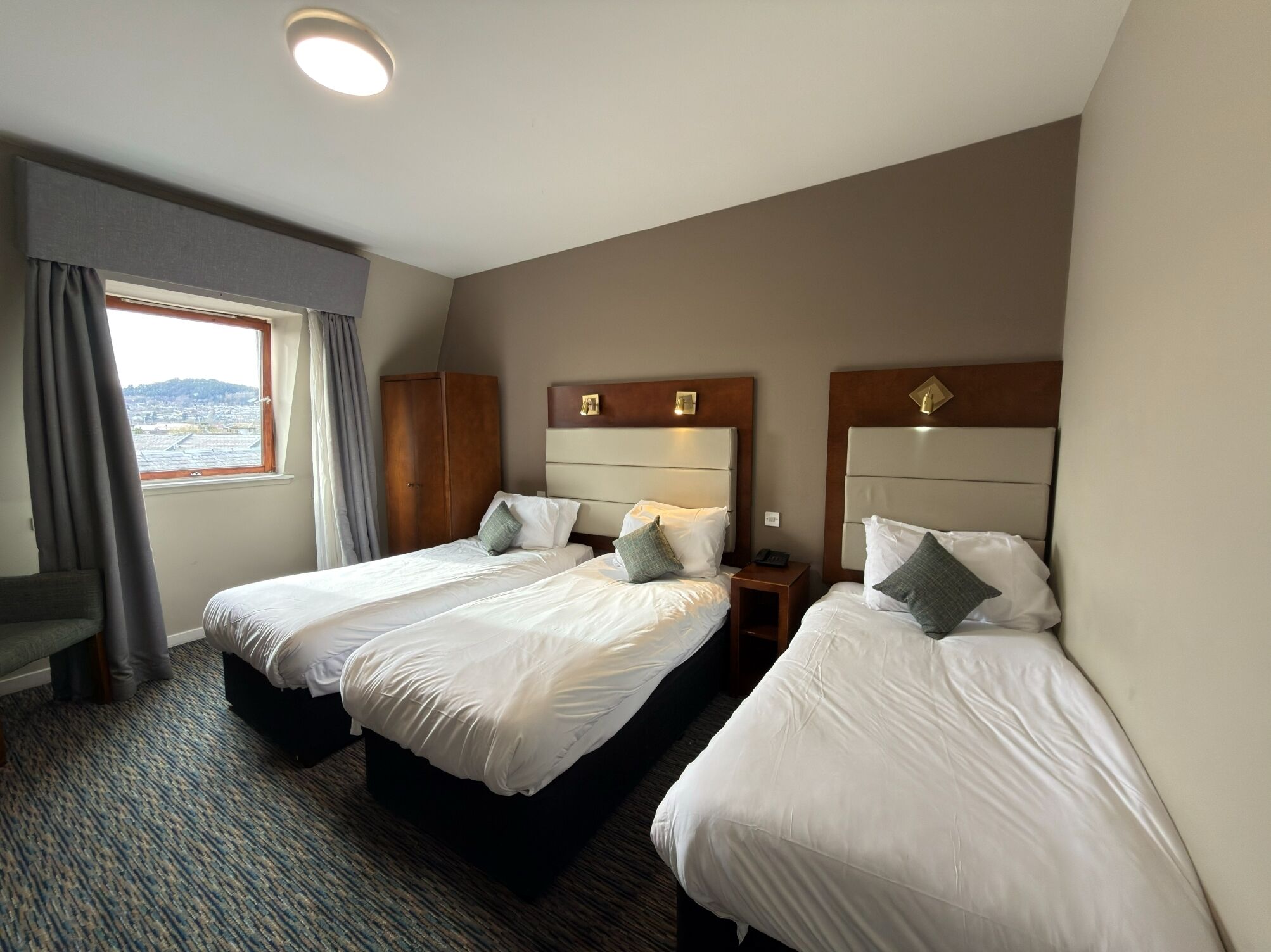 Habitación de hotel con dos camas individuales, un escritorio pequeño, una ventana con cortinas y vista a un paisaje urbano.