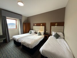 Habitación de hotel con dos camas individuales, un escritorio pequeño, una ventana con cortinas y vista a un paisaje urbano.