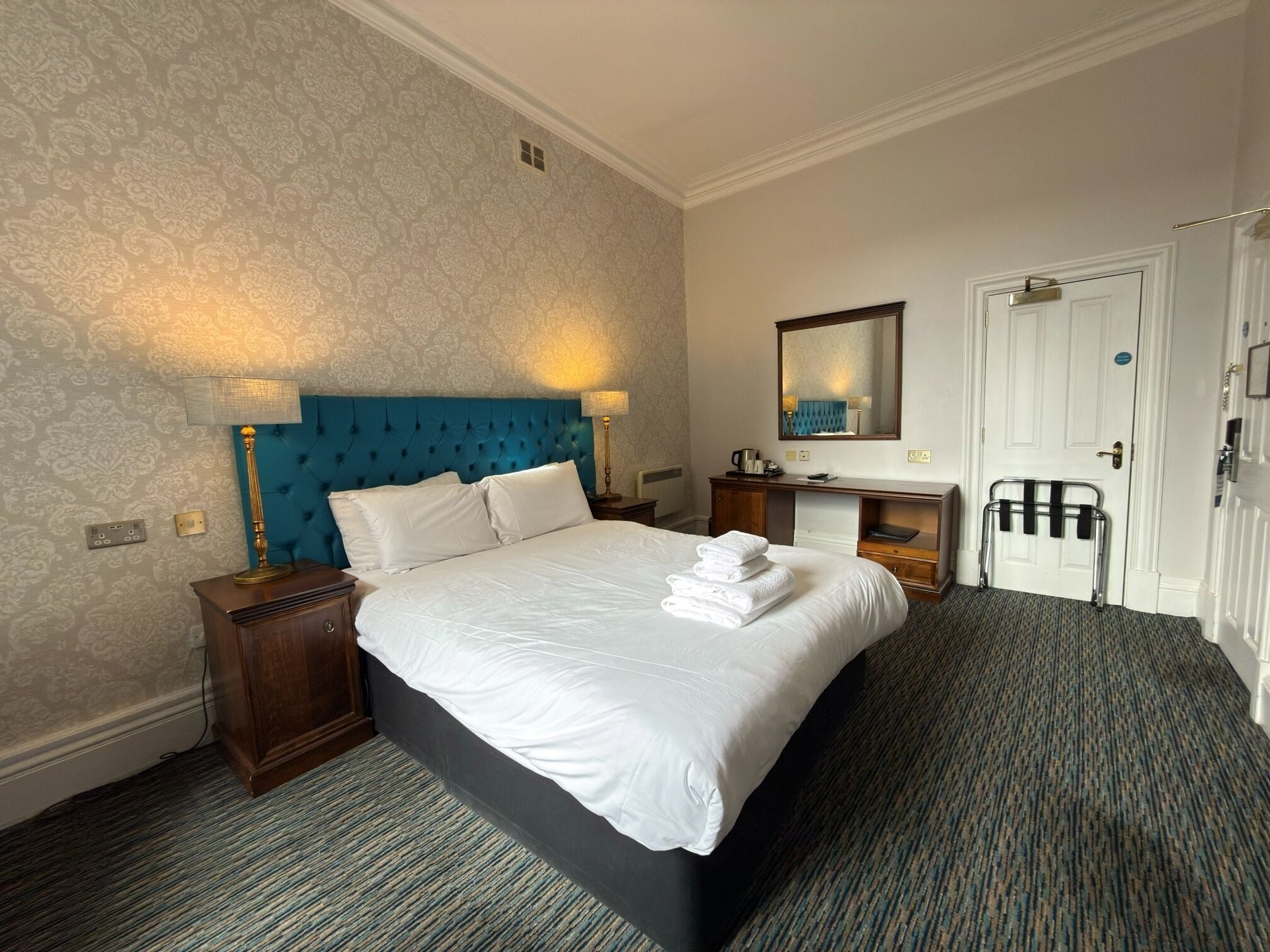 A hotel room with a large bed, a bedside table, a dresser, a mirror, and a door.