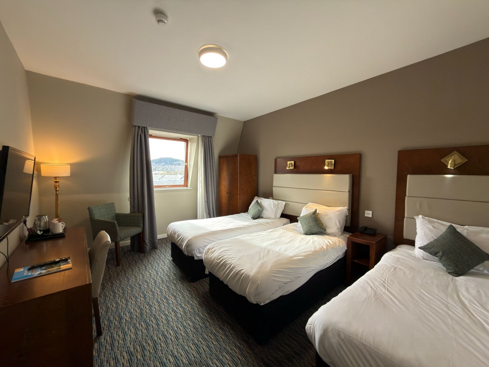 A hotel room with two beds, a desk, a chair, a lamp, and a window with curtains.