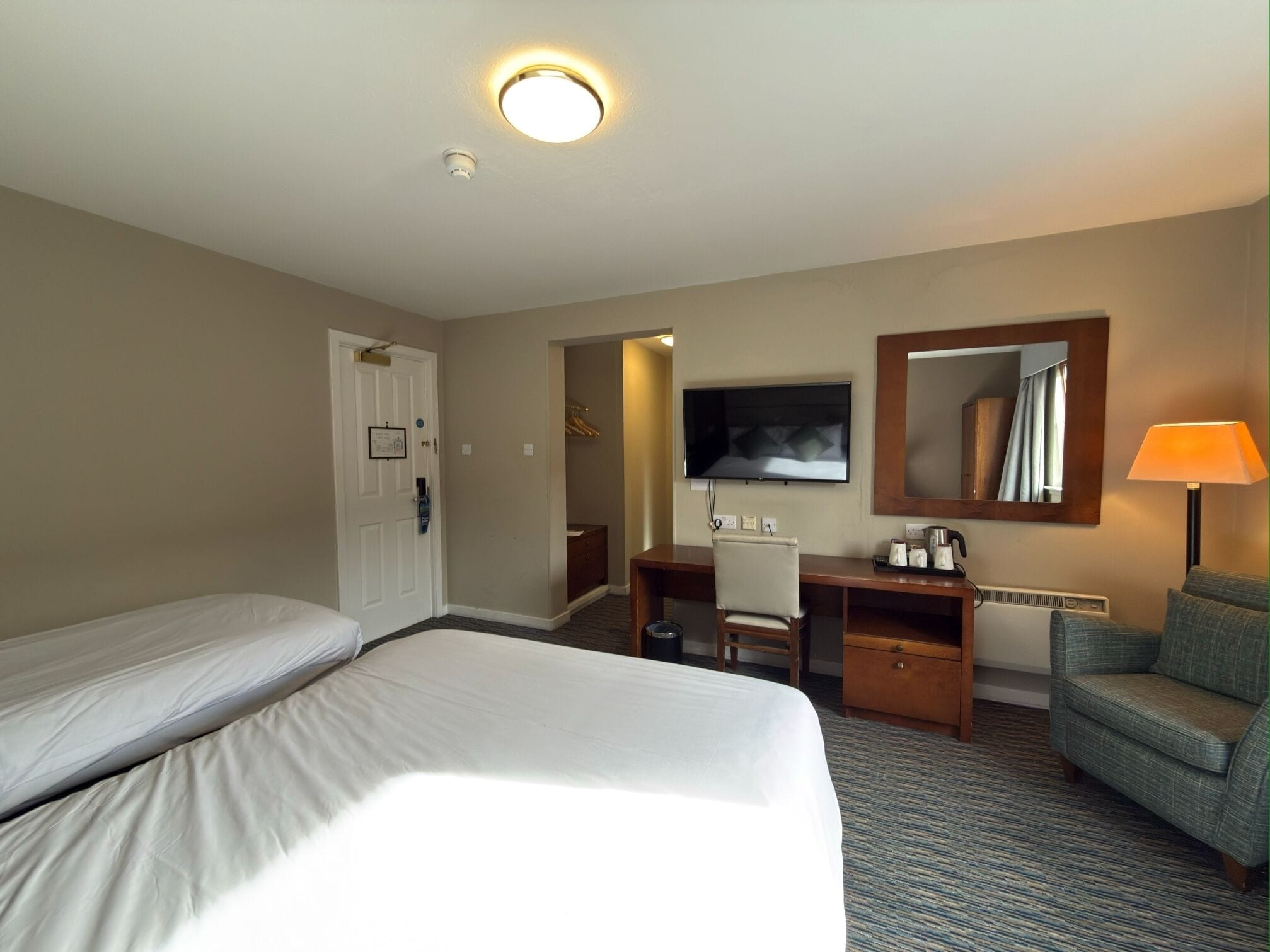 A hotel room with two beds, a desk, a chair, a TV, a mirror, and a lamp.