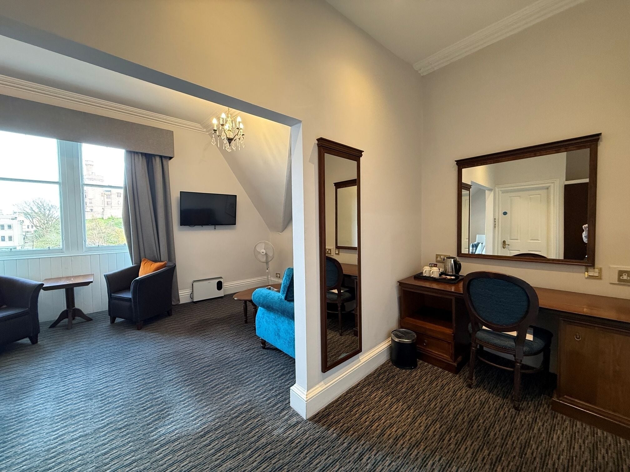 Habitación de hotel con escritorio, dos sillas, sofá, televisión, espejo y ventana con vistas a edificios.