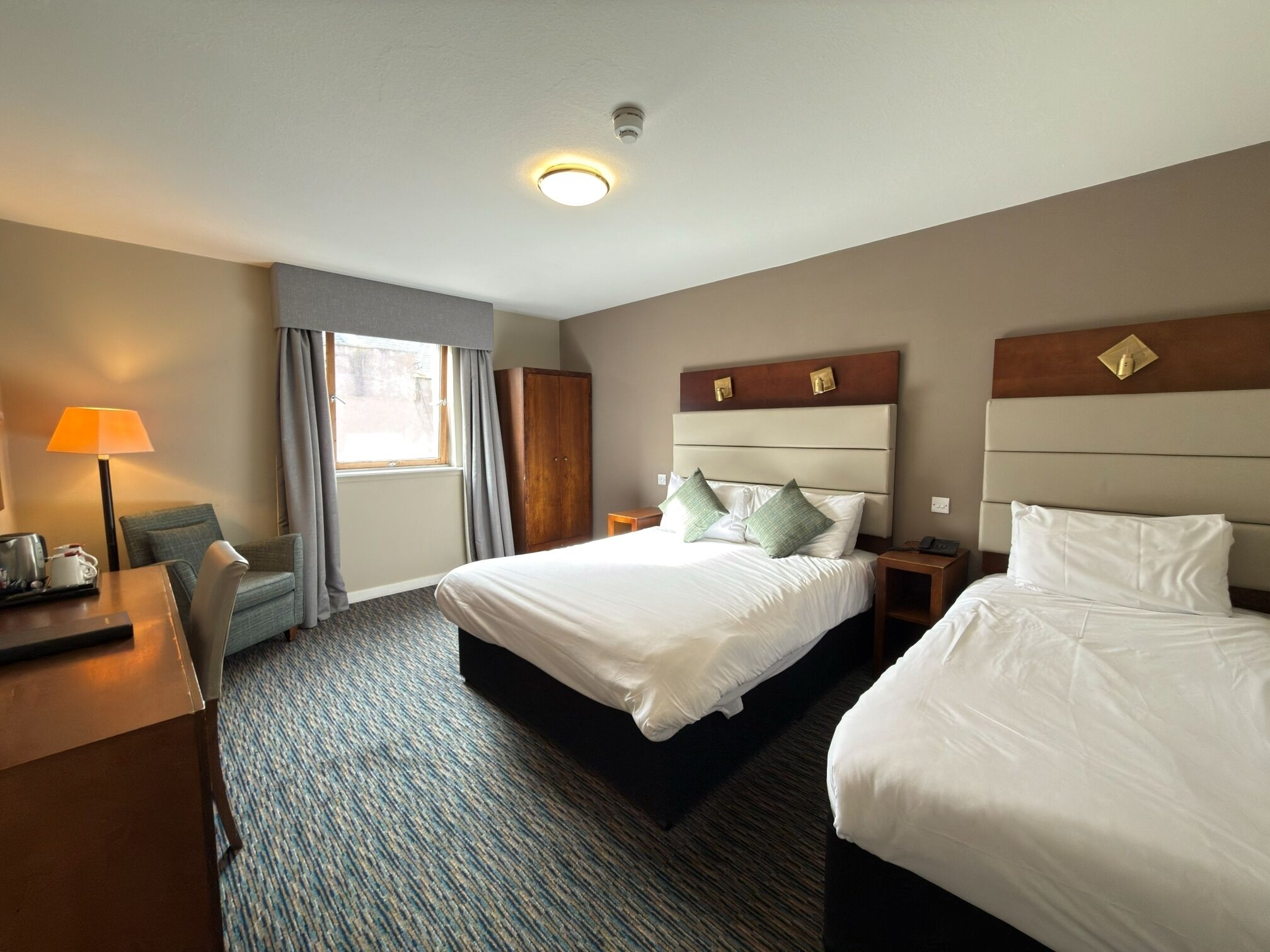 A hotel room with two beds, a desk, a lamp, and a window with curtains.