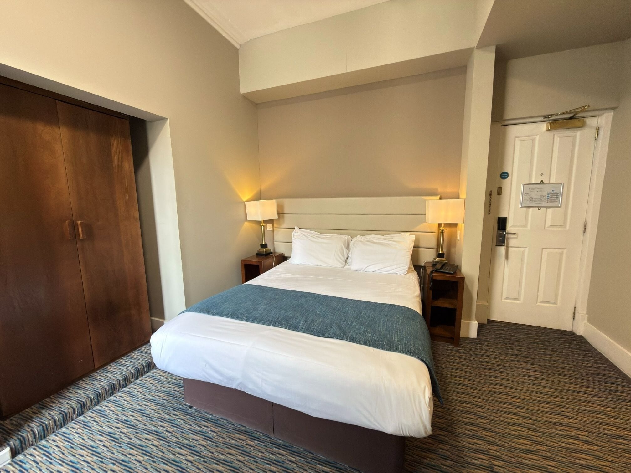 A hotel room with a large bed, two bedside tables, a wooden wardrobe, and a door with a sign.