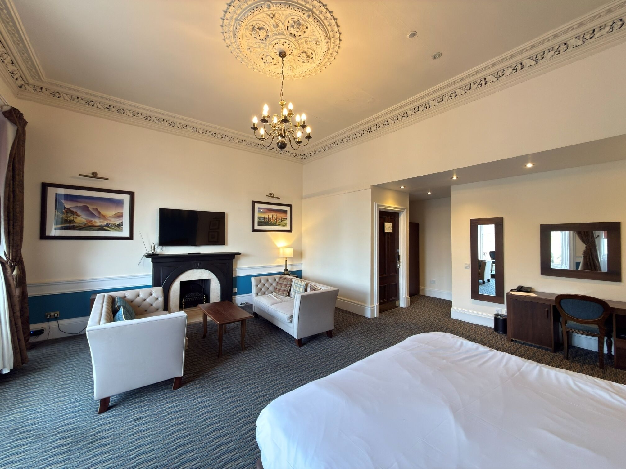 A hotel room with a large bed, a sofa, a TV, a fireplace, and a chandelier.