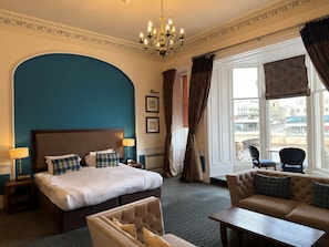 A hotel room with a large bed, a sofa, a table, and a view of the city through the window. - Columba Hotel Inverness by Compass Hospitality (Inverness)