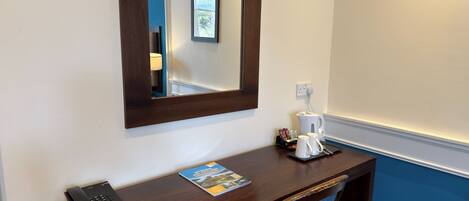 A hotel room with a wooden desk, a telephone, a coffee maker, a magazine, and a mirror on the wall.