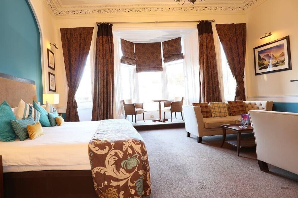 A hotel room with a bed, a sofa, a small table, and a chair. There are curtains, a picture on the wall, and a window with a view.