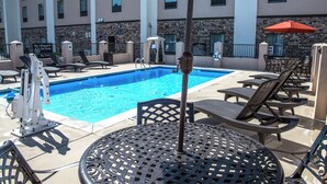 Zona de piscina con sillas de descanso y una mesa, señal de socorrista, y un edificio con fachada de piedra y ventanas.