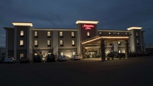 Un hotel Hampton Inn con diseño moderno, que presenta un llamativo letrero de neón, un acceso cubierto y un estacionamiento con varios autos.