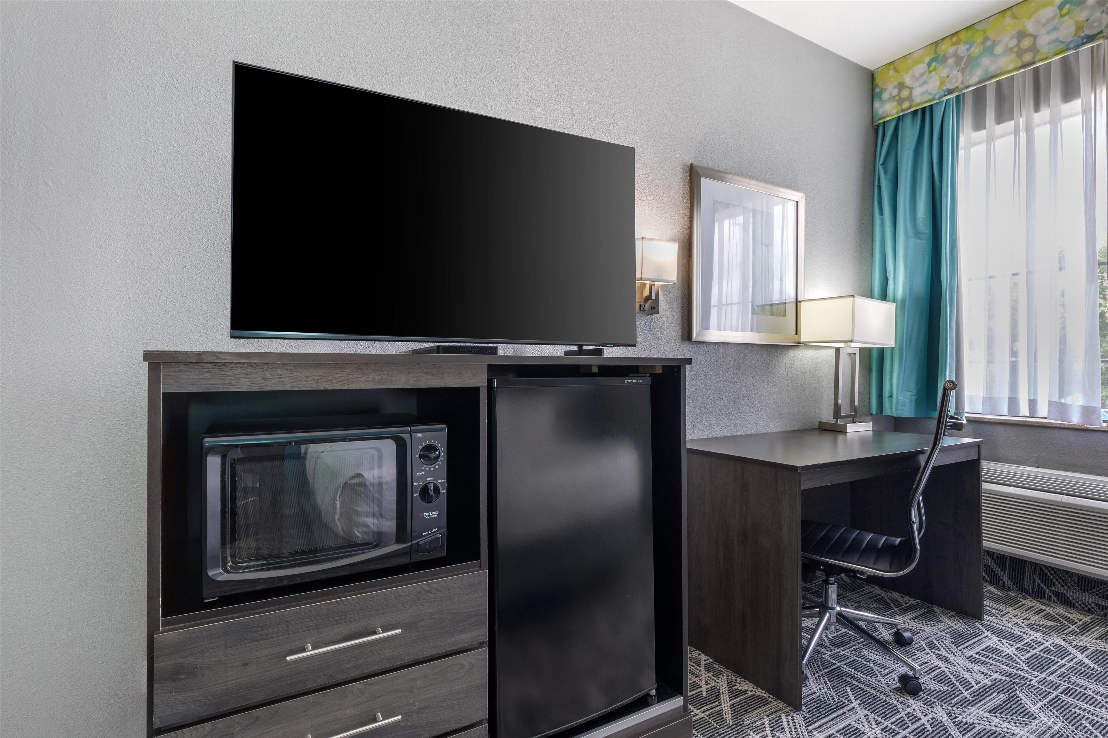 A compact hotel room with a flat-screen TV, a built-in microwave, a small refrigerator, a desk with a chair, and a window with curtains.