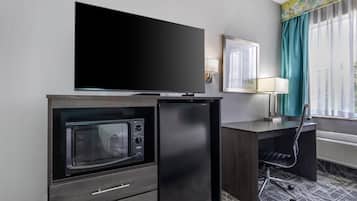 A compact hotel room with a flat-screen TV, a built-in microwave, a small refrigerator, a desk with a chair, and a window with curtains.