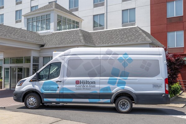 Un fourgon blanc orné de motifs en losanges bleus et portant l’inscription « Hilton Garden Inn Queens / Aéroport JFK » est garé devant un immeuble doté de grandes fenêtres et d’une façade en briques rouges.