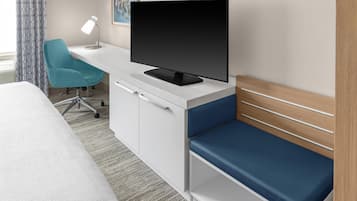 A hotel room with a bed, a desk with a television, a chair, and a desk lamp.