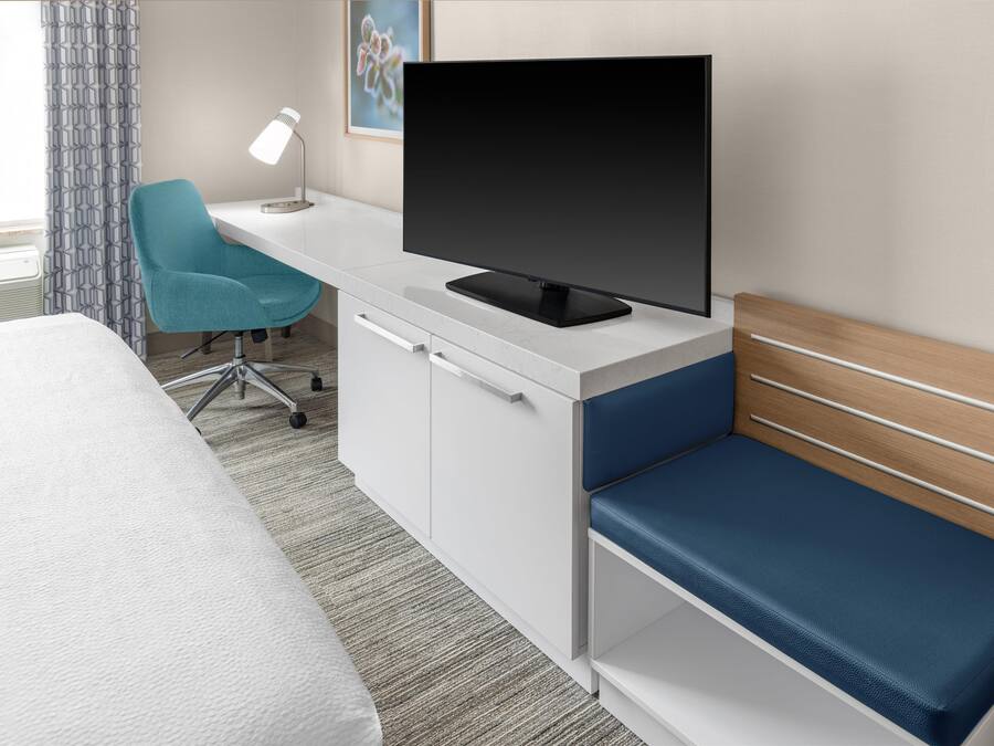 Quarto de hotel com cama, escrivaninha com televisão, cadeira e luminária.