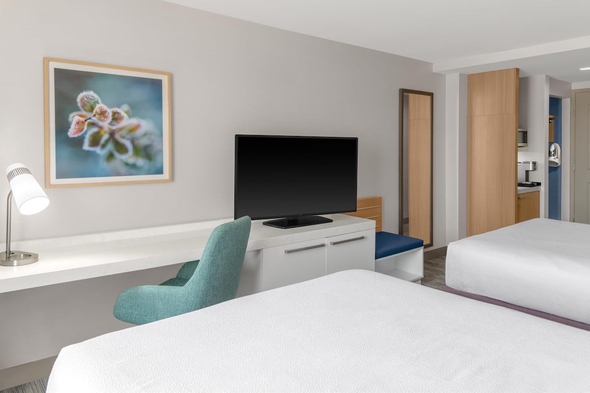 A hotel room with a bed, a desk with a TV, a chair, and a painting on the wall.