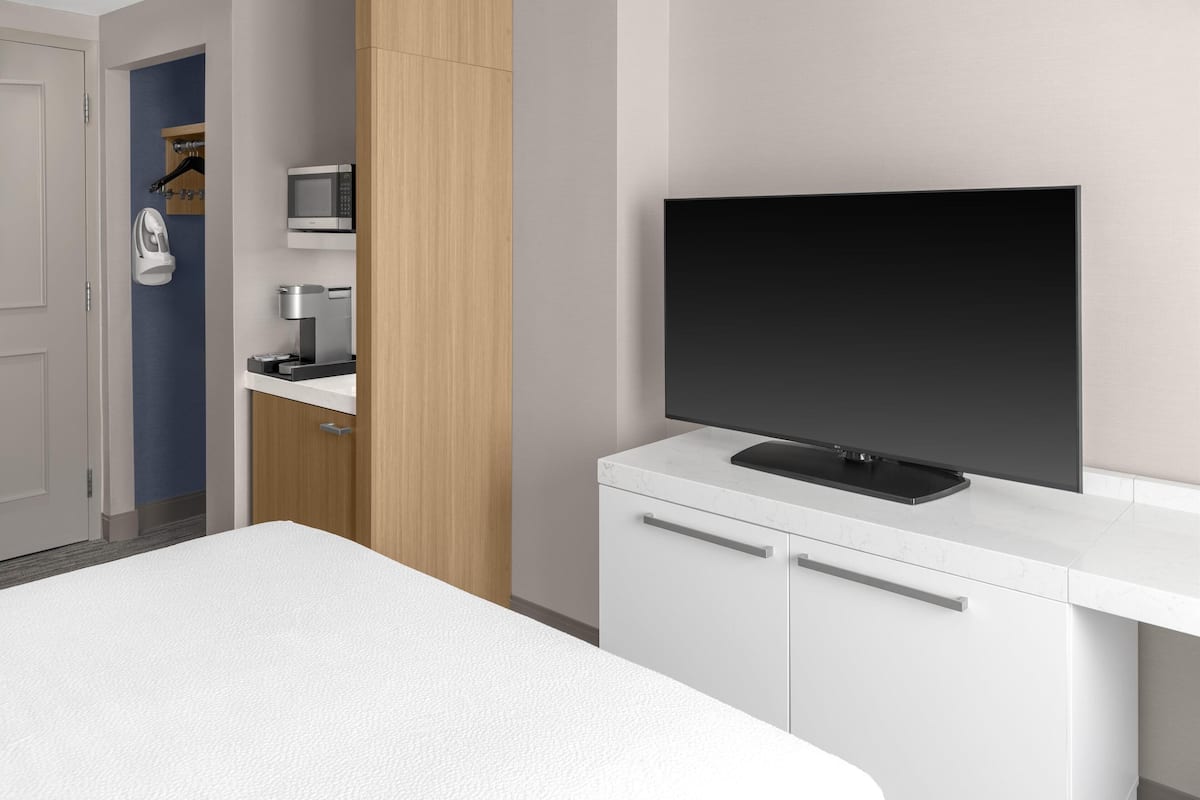 Una habitación de hotel moderna con una cama, un televisor de pantalla plana montado en un mueble blanco, un microondas y una cafetera.