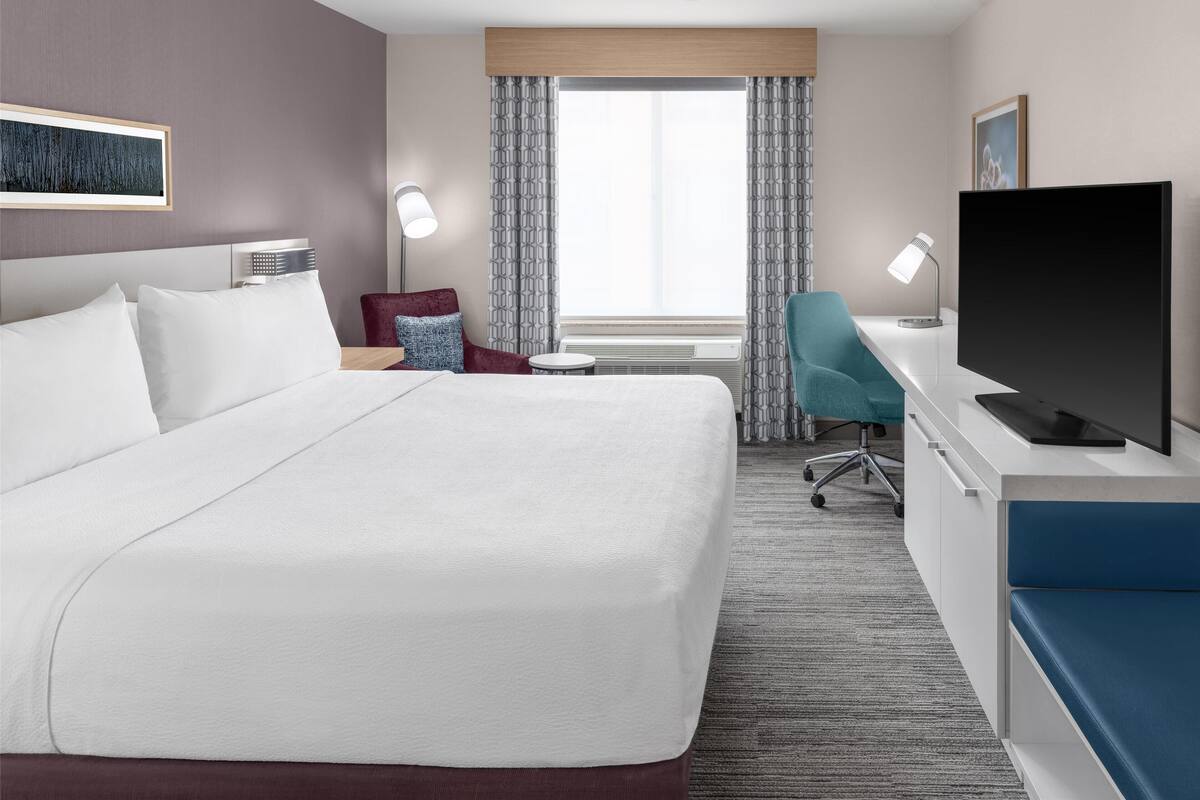 A hotel room with a large bed, a desk with a flat-screen TV, a chair, a small table, and a window with curtains.