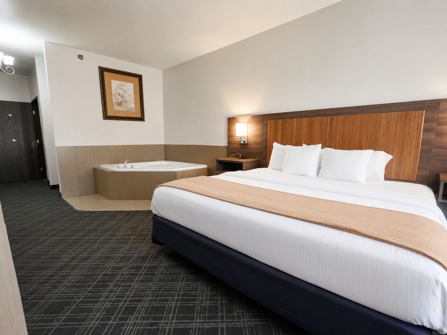 Une chambre d’hôtel avec un grand lit, une salle de bain séparée avec une baignoire, une tête de lit en bois et un tableau encadré au mur.