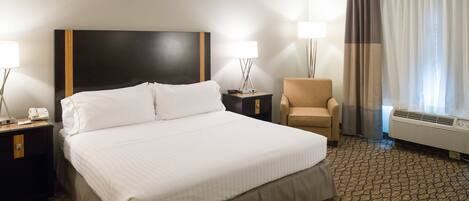 A hotel room with a large bed, a chair, a nightstand with a lamp, a bedside table, and a window with curtains.