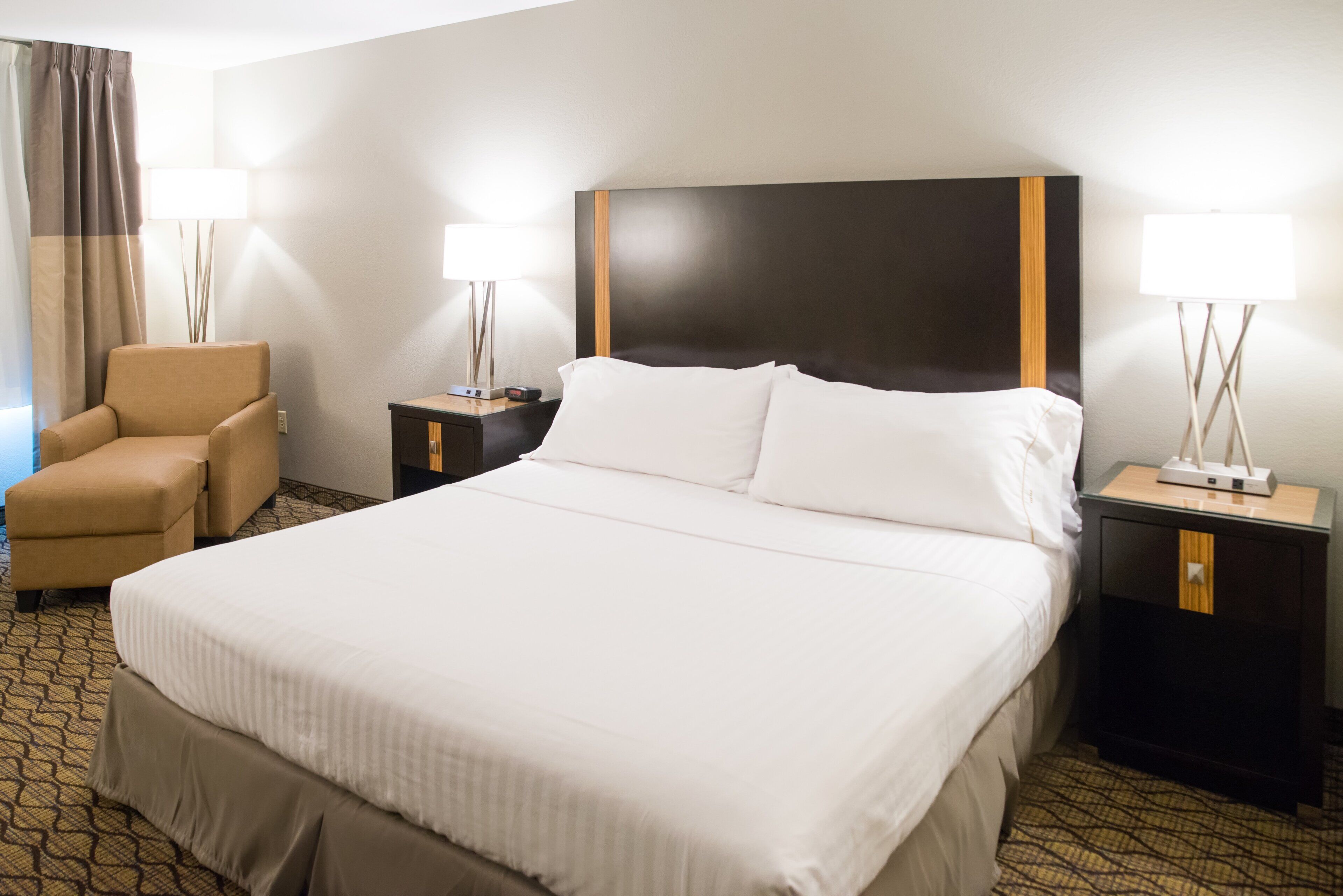 Una habitación de hotel con una cama grande, dos mesitas de noche, una silla y una lámpara.
