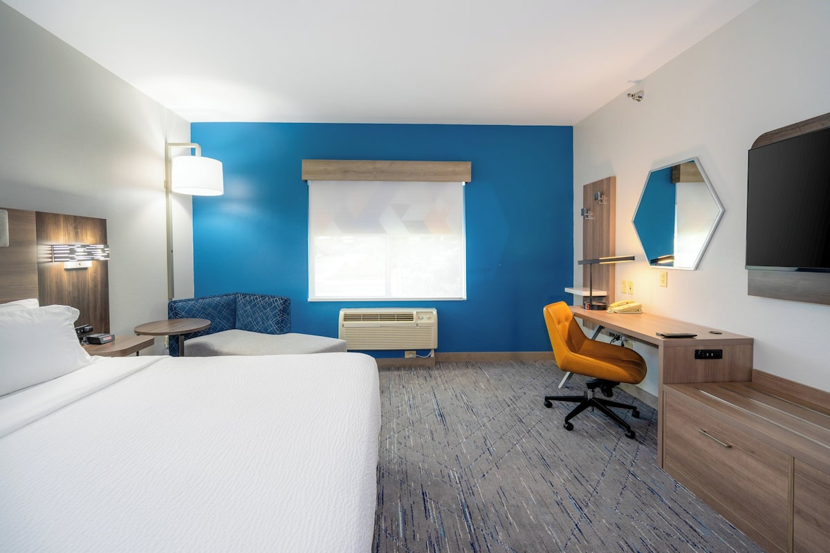 A hotel room with a bed, a desk, a chair, a window with blinds, a heater, and a mirror on the wall.