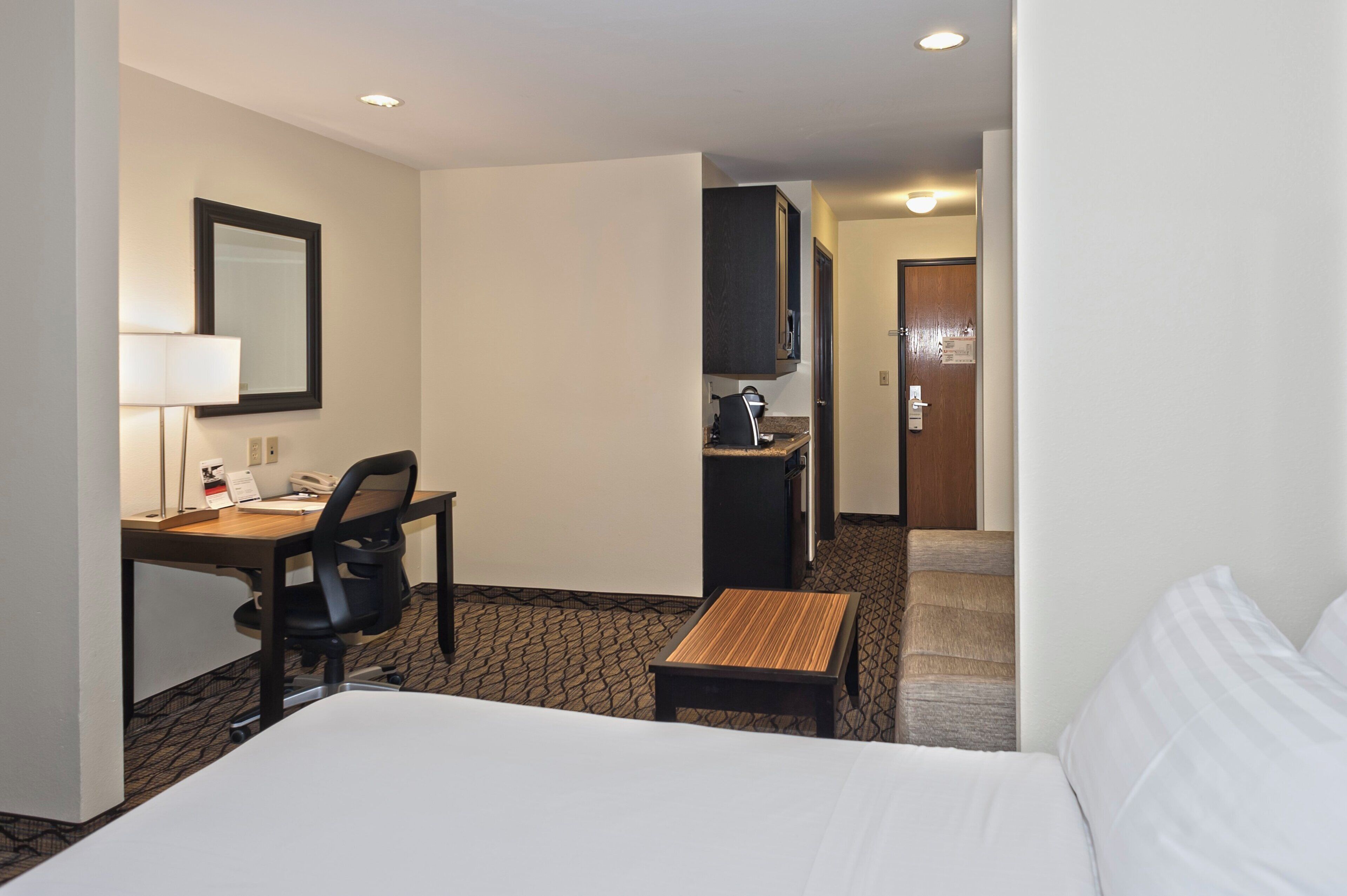 A hotel room with a bed, a desk, a chair, a small table, a mirror, a television, and a door leading to a bathroom.