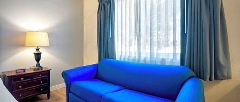 A room with a blue sofa, a wooden nightstand with a lamp, and a window with curtains.