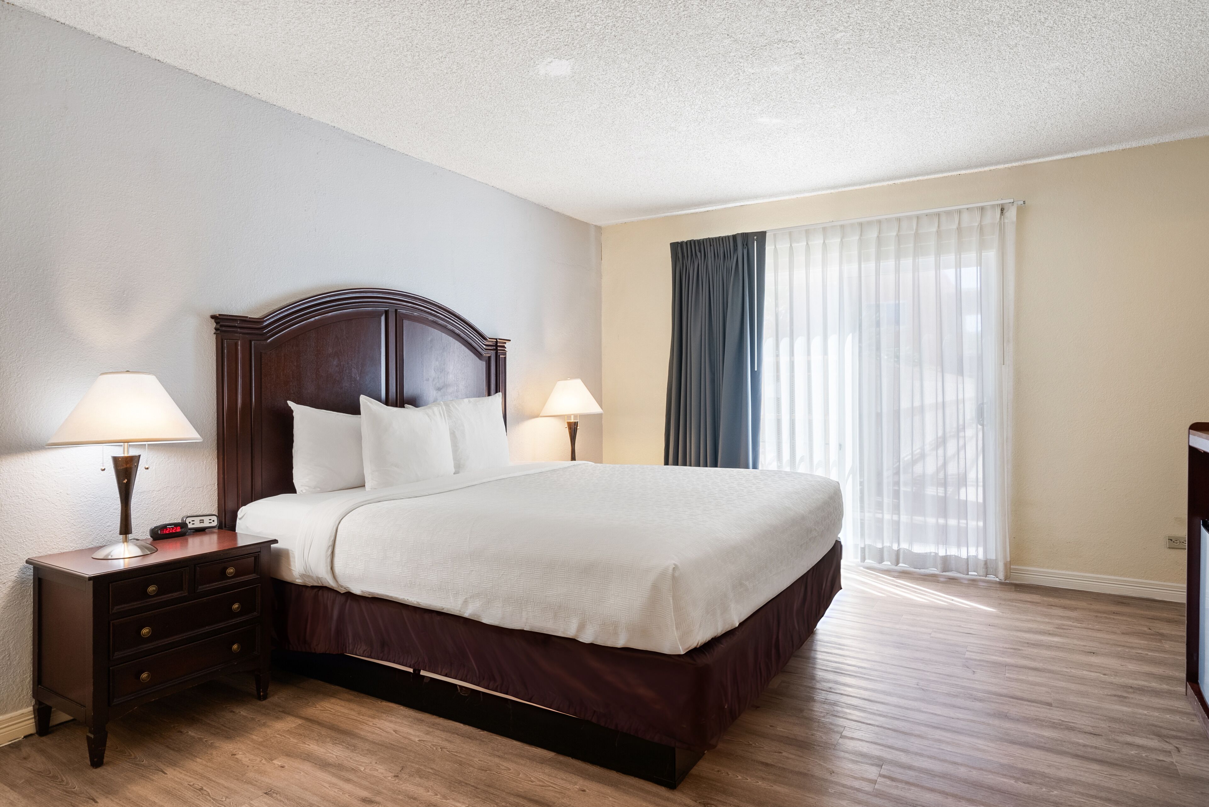 A neatly made bed with a wooden headboard, two bedside tables with lamps, a large window with sheer curtains, and a wooden floor.