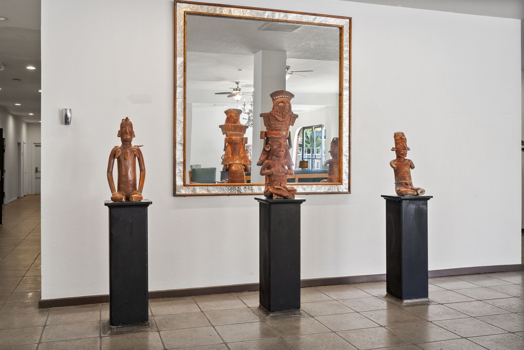 Three wooden sculptures on pedestals in a hallway with a large mirror on the wall.