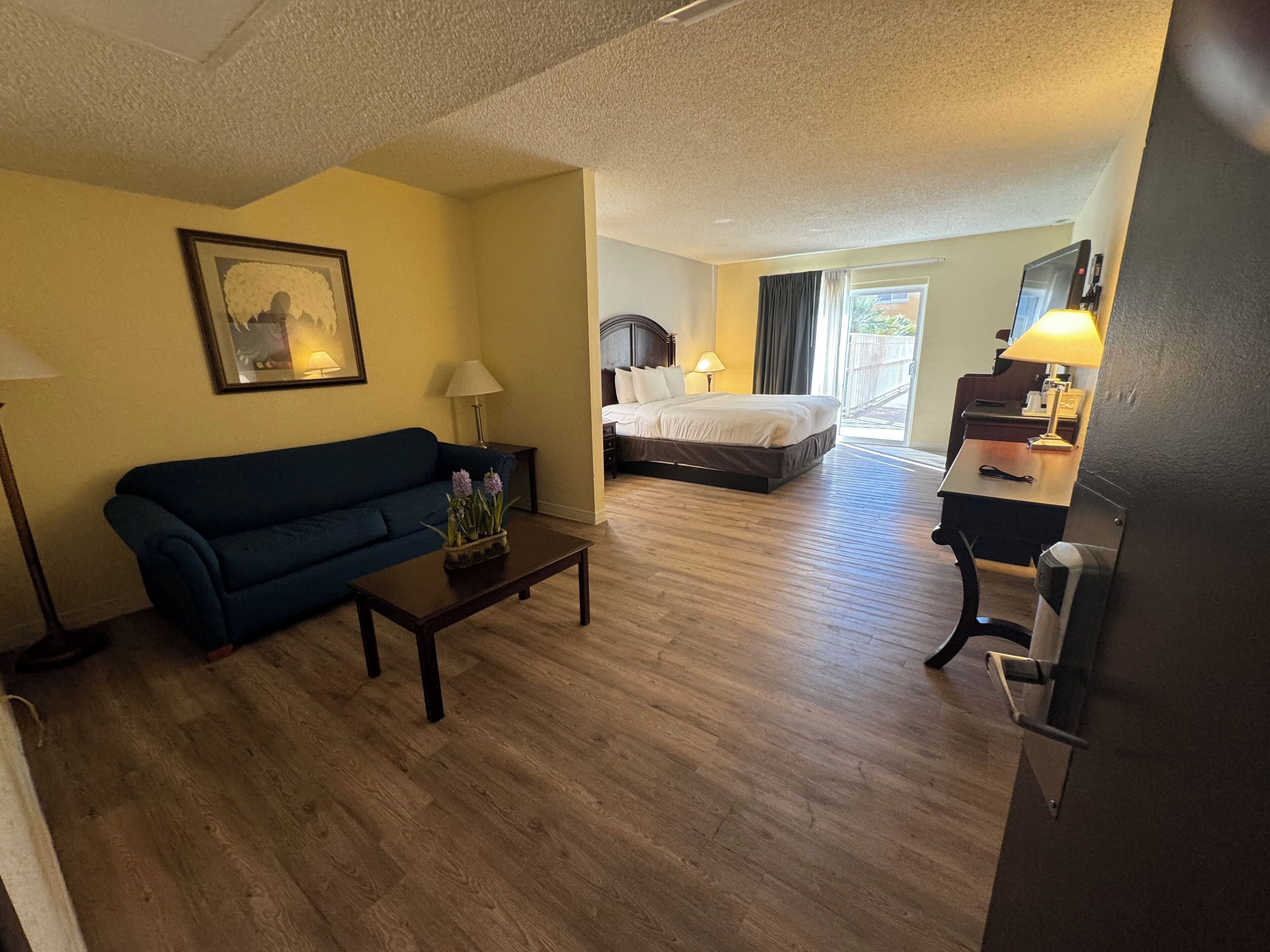A hotel room with a bed, a sofa, a coffee table with flowers, a desk with a lamp, and a painting on the wall.