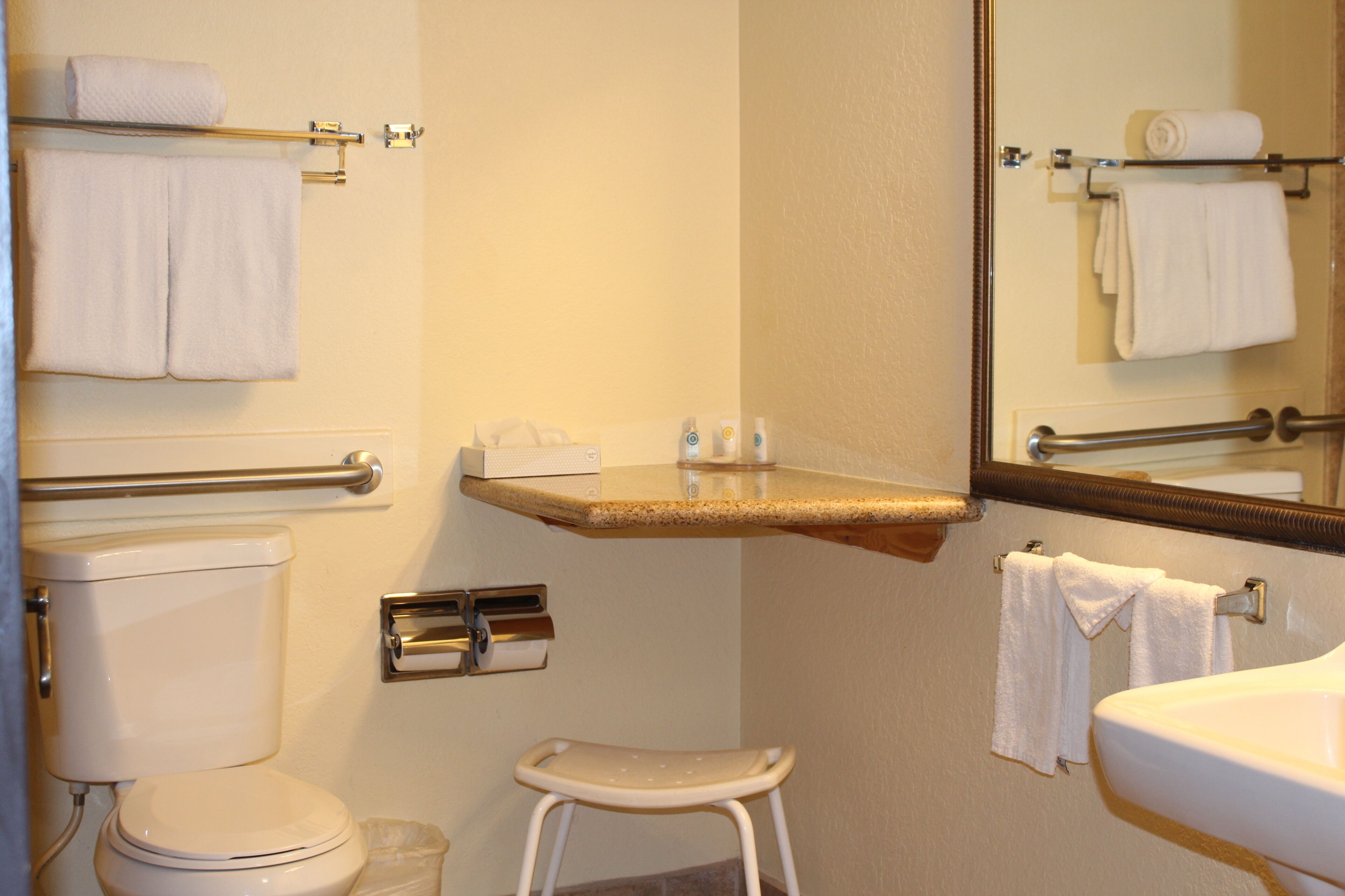 Un baño con inodoro, lavamanos y un taburete pequeño. Hay toallas en la pared y un espejo sobre el lavamanos.